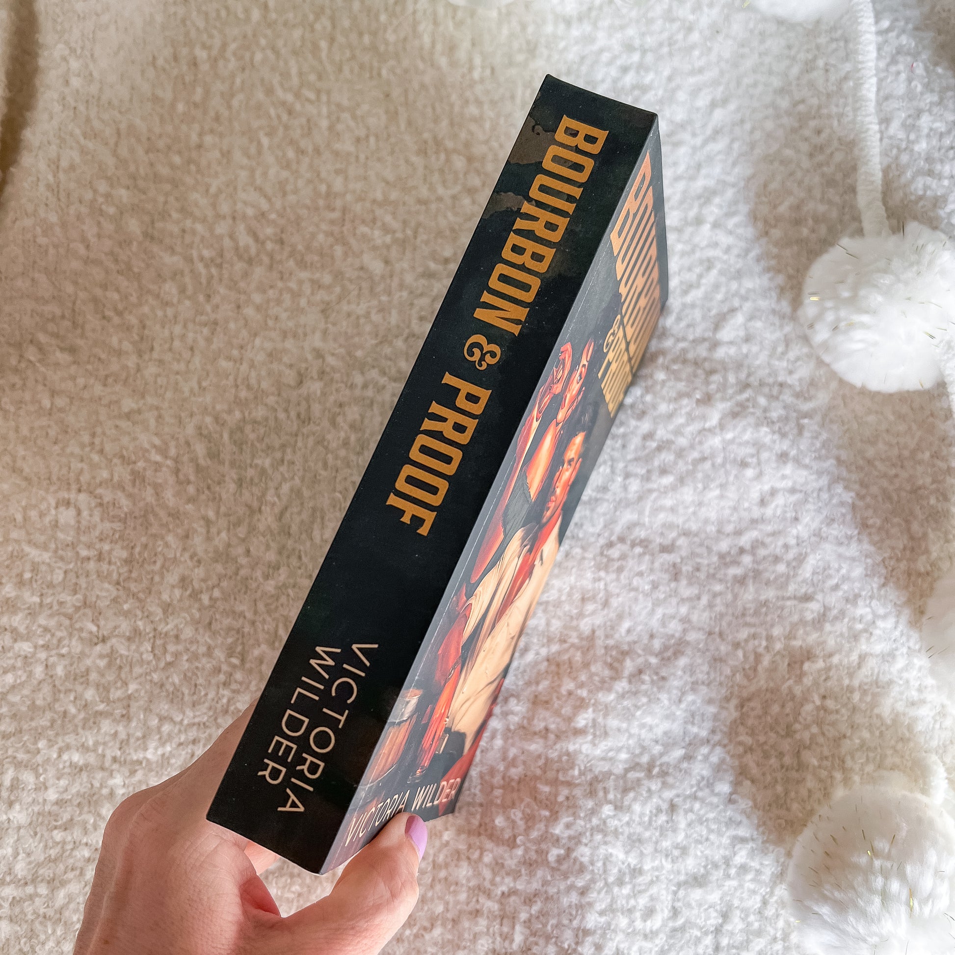 A hand holds a book titled 'Bourbon & Proof' by Victoria Wilder, showing the black spine with gold lettering, against a soft beige textured blanket background.