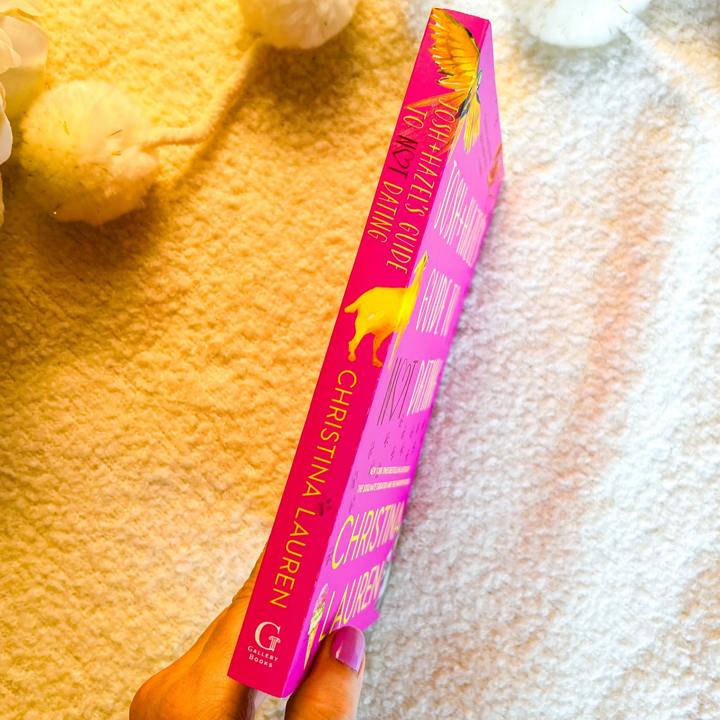 A hand holds a bright pink book by Christina Lauren, showing the spine with gold lettering and a yellow bird illustration, against a textured cream background with soft decorative flowers.