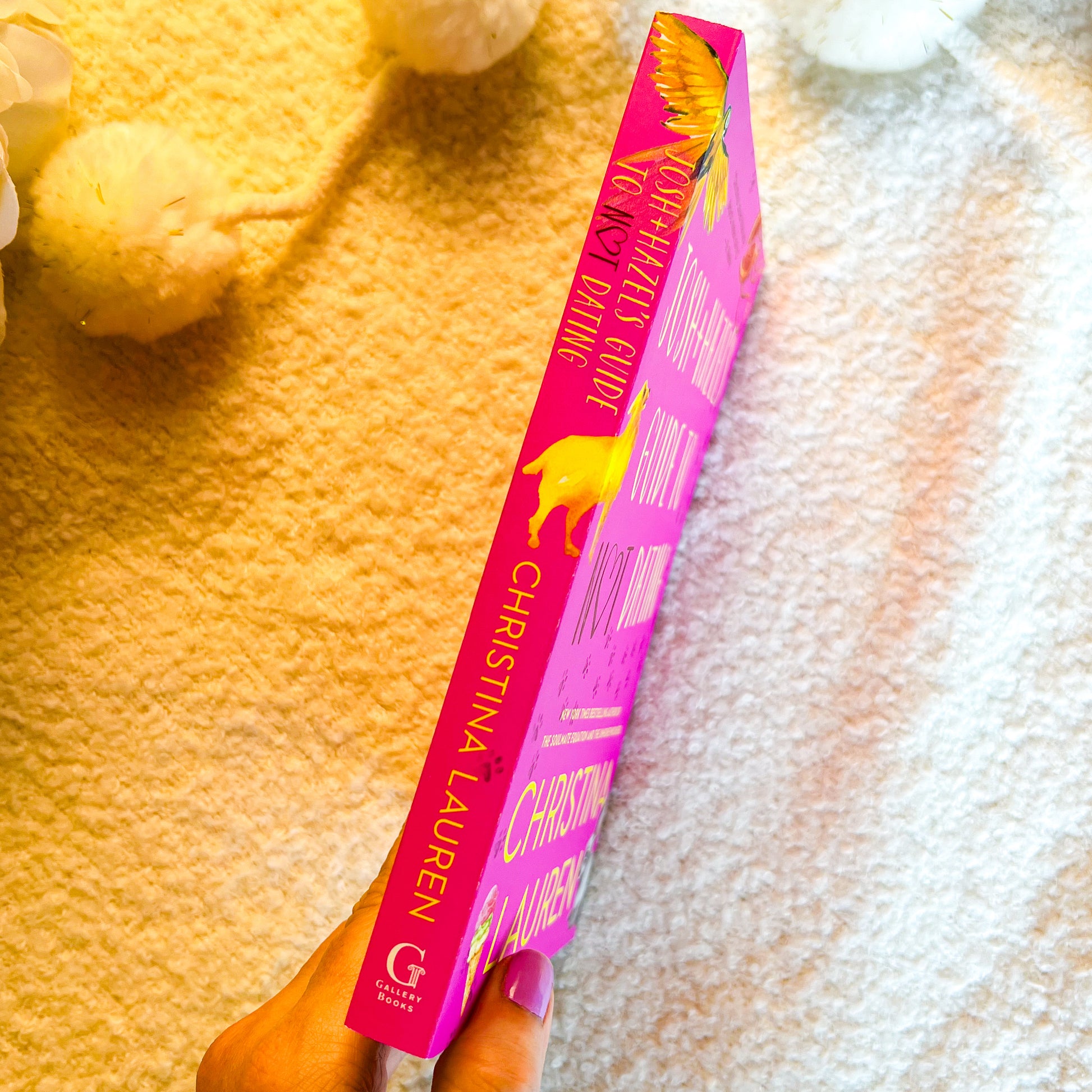 A hand holds a bright pink book by Christina Lauren, showing the spine with gold lettering and a yellow bird illustration, against a textured cream background with soft decorative flowers.