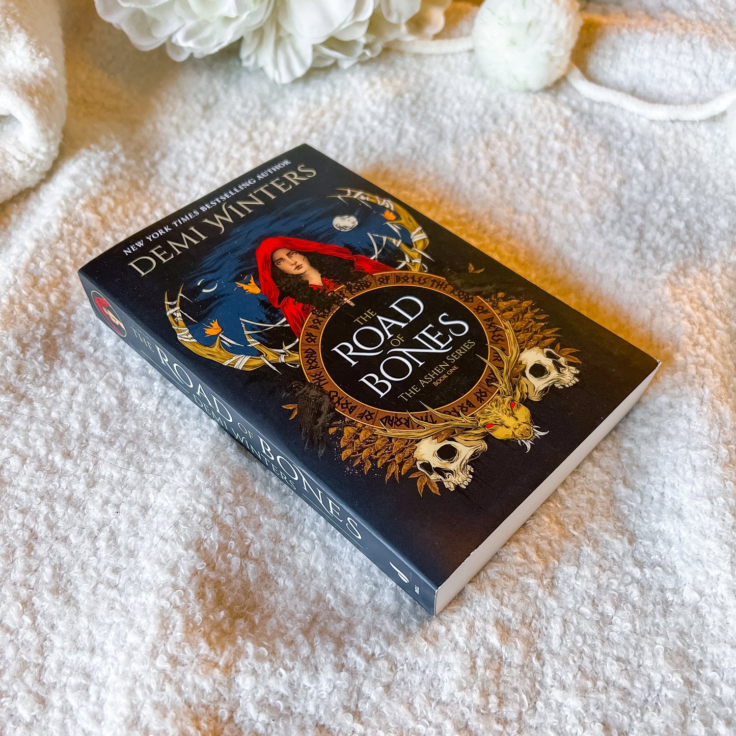 Book titled 'The Road of Bones' by Demi Winter on a textured surface with flowers.