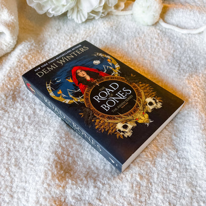 Book titled 'The Road of Bones' by Demi Winter on a textured surface with flowers.