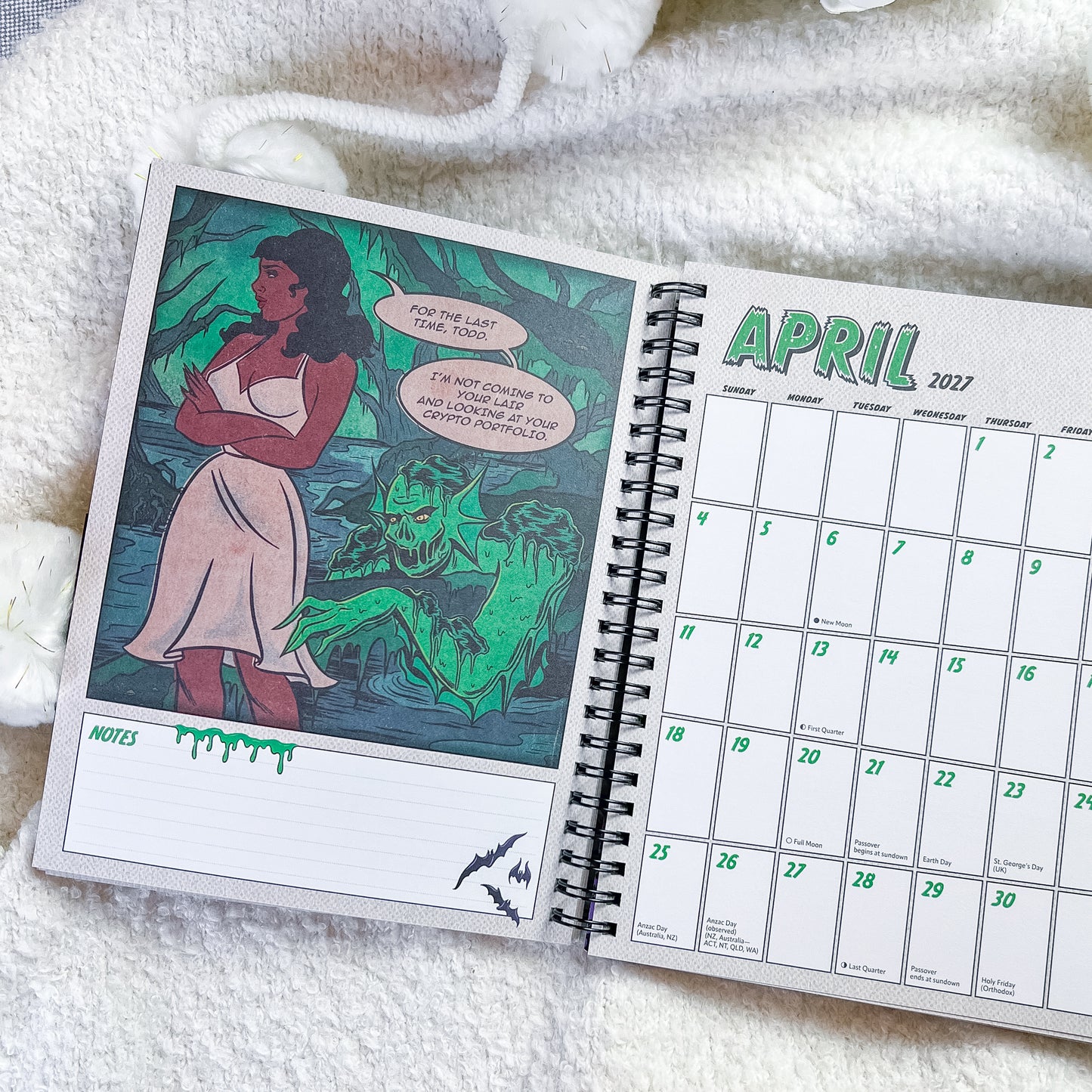 An open spiral-bound planner or calendar laid on a soft white textured blanket. The left page shows a comic-style illustration with a woman in a white dress and green monster artwork, and the right page shows the April 2027 calendar grid with lined notes section at the bottom.