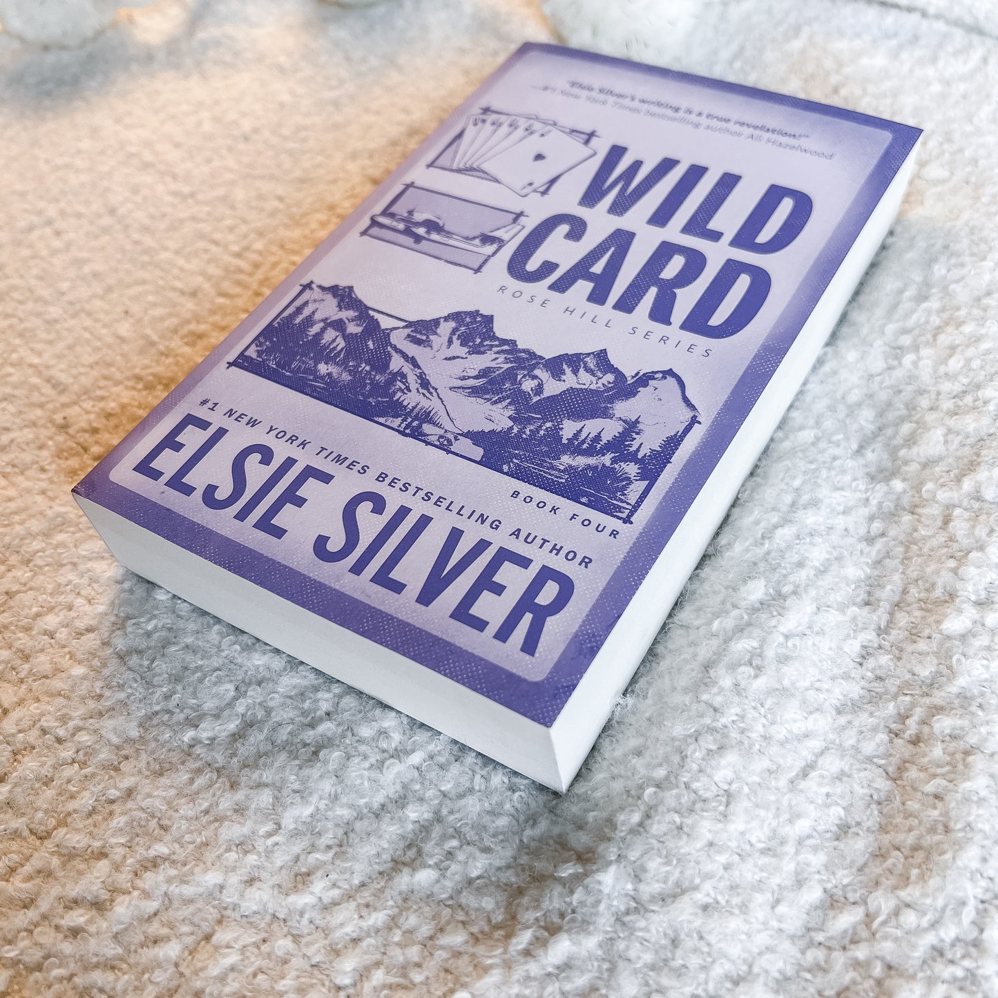 A paperback copy of the book 'Wild Card' by Elsie Silver, featuring a purple and white cover with mountain illustrations, resting on a textured beige carpet.