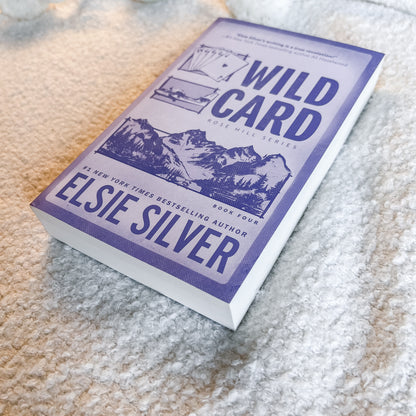 A paperback copy of the book 'Wild Card' by Elsie Silver, featuring a purple and white cover with mountain illustrations, resting on a textured beige carpet.