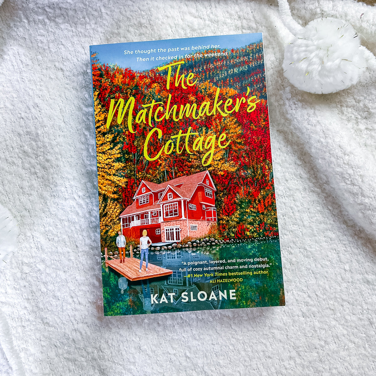 The book cover of 'The Matchmaker's Cottage' by Kat Sloane is displayed flat on a white textured blanket, with a white knitted sock in the top right corner.