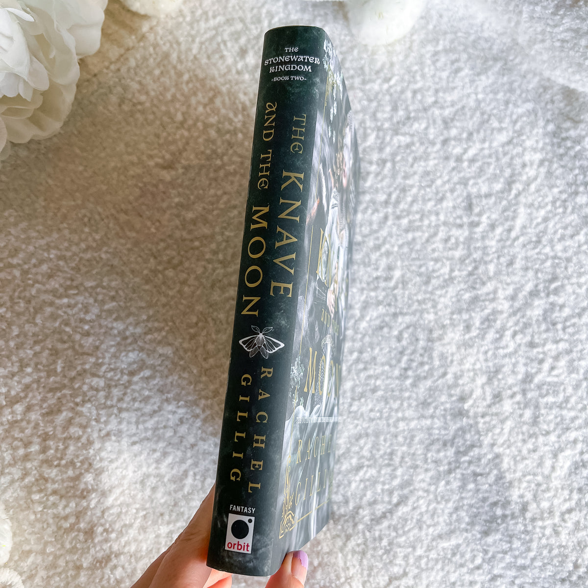 A hand holds a hardcover book titled 'The Knave and the Moon' by Rachel Gillig, showing the spine with gold lettering on a dark background, against a soft white textured blanket backdrop.