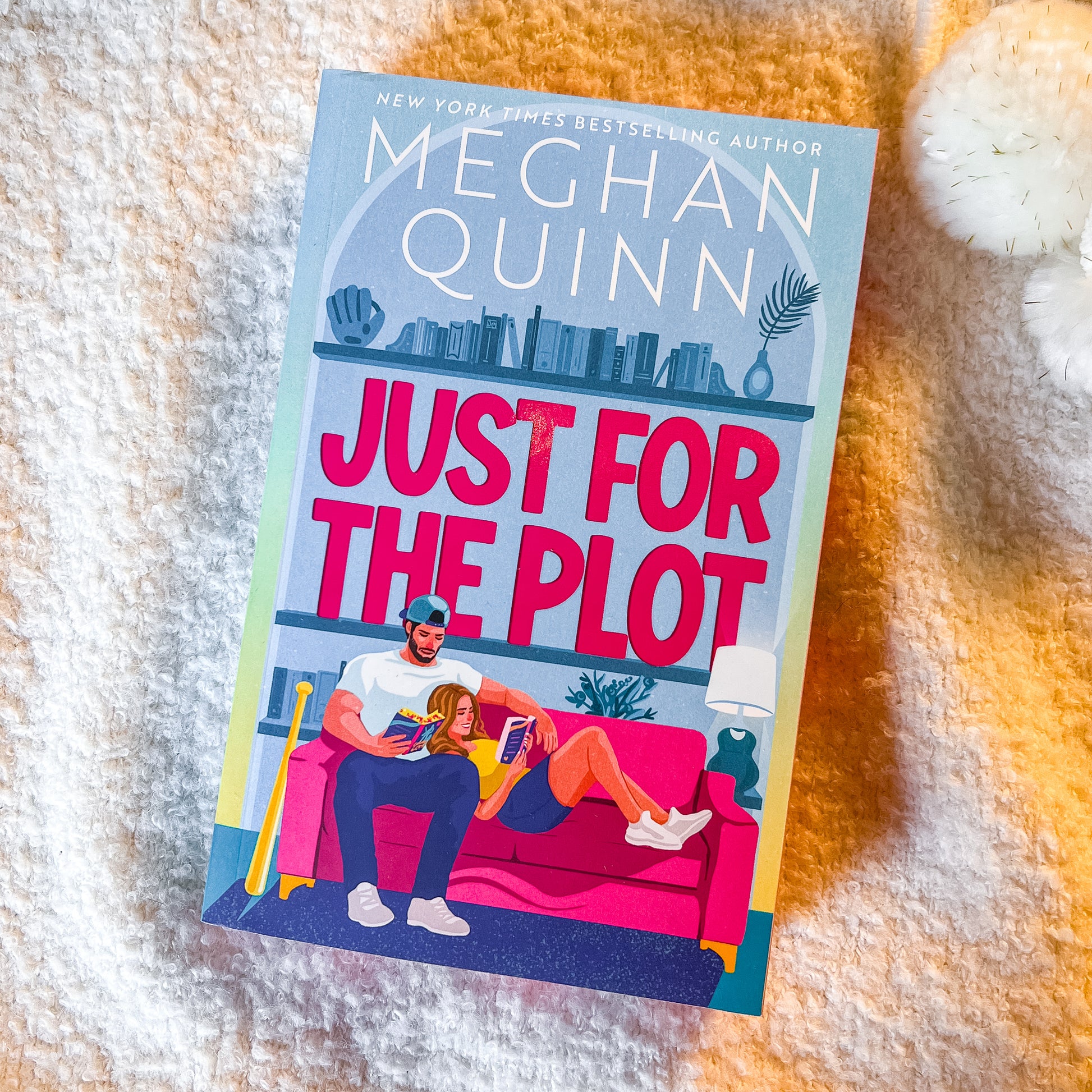 A paperback copy of the book 'Just for the Plot' by Meghan Quinn lies on a textured cream-colored blanket. The cover features a colorful illustration of a man and a woman sitting on a pink couch in front of bookshelves.