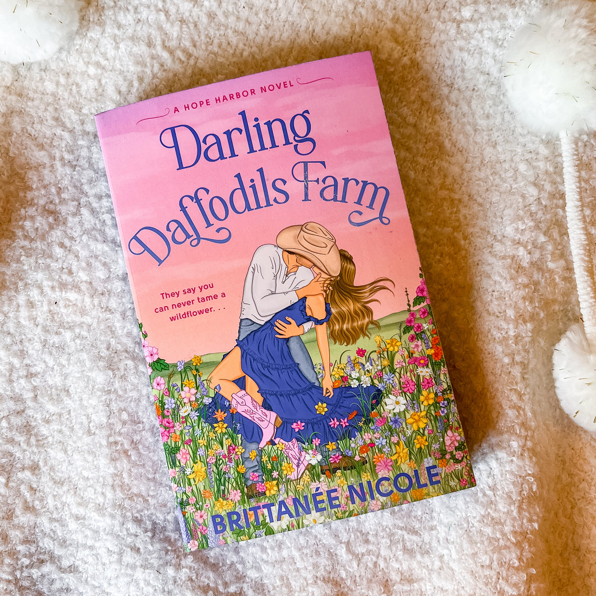 A paperback book titled "Darling Daffodils Farm" by Brittanie Nicole, featuring a pink cover with an illustration of a woman in a blue dress and cowboy hat embracing a man in a flower field, photographed on a soft white textured blanket.
