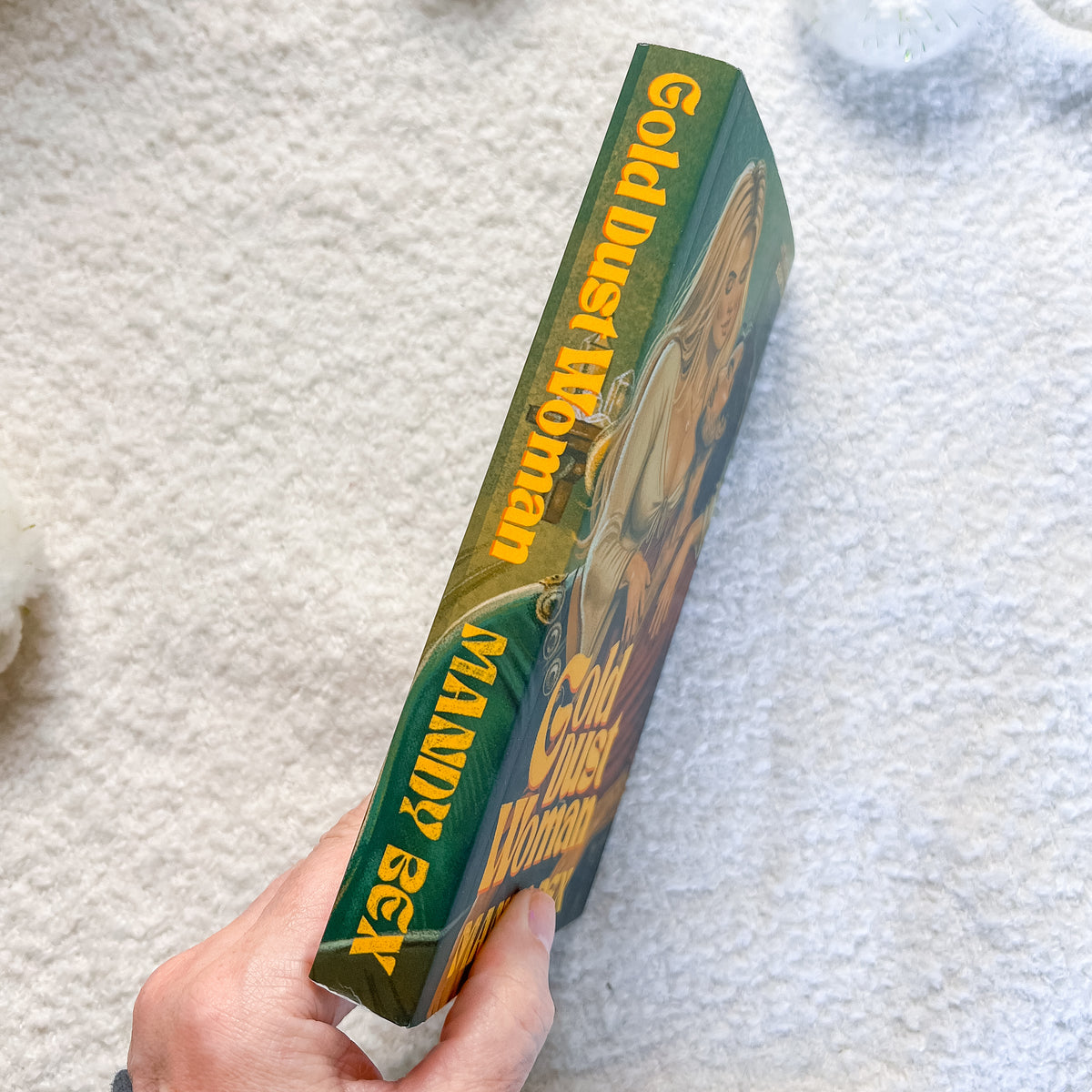 A hand holds a book titled 'Gold Dust Woman' by Mandy Bex, showing the green and gold spine, against a white textured background.
