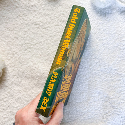 A hand holds a book titled 'Gold Dust Woman' by Mandy Bex, showing the green and gold spine, against a white textured background.