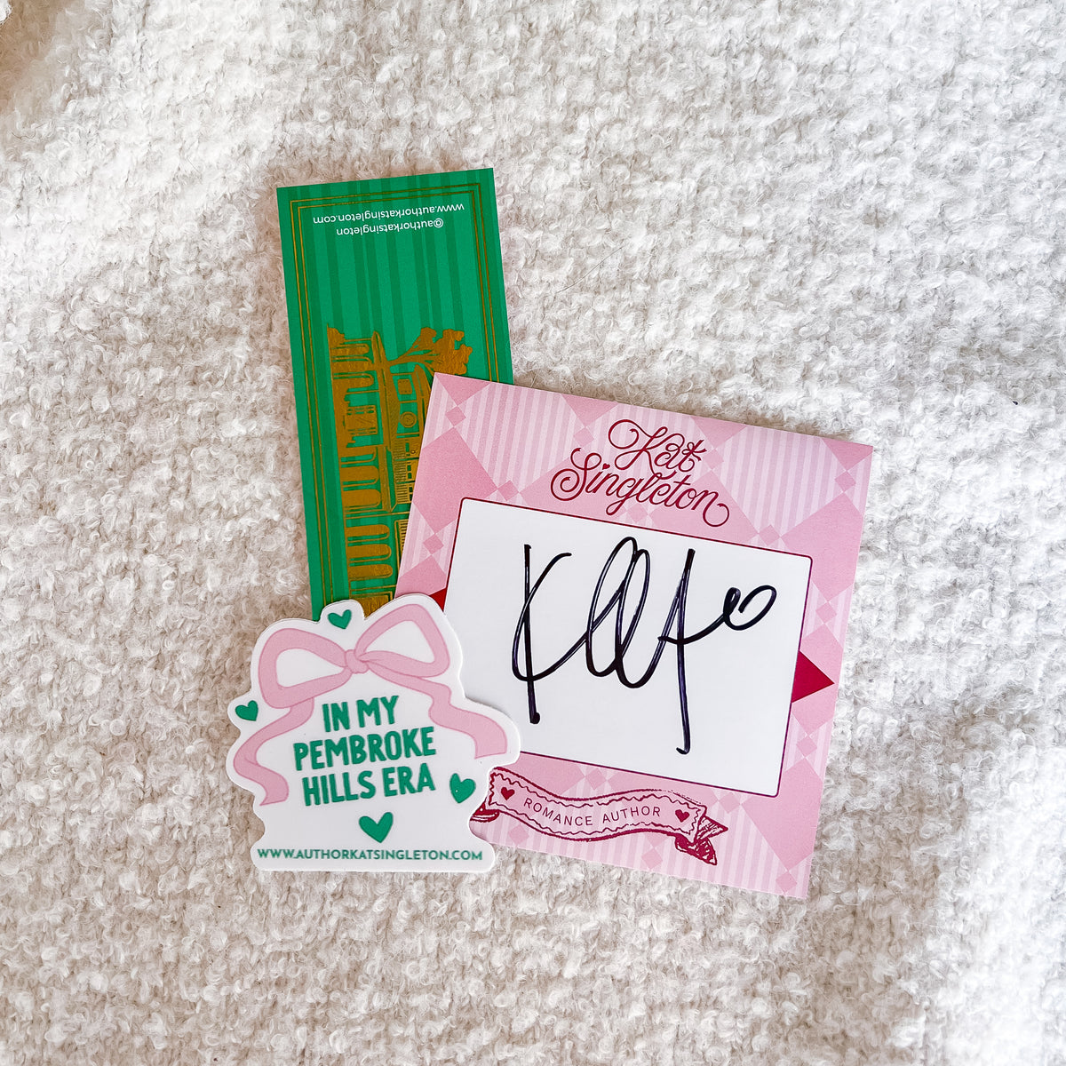 A pink and white autographed 'All Is Singled' card with a black ink signature, displayed alongside a pink 'In My Pembroke Hills Era' bow-shaped sticker and a green collectible card on a white textured rug.