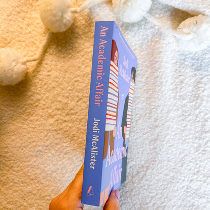 A hand holds a book titled 'An Academic Affair' by Jodi McAlister. The book cover is predominantly blue with red and white text and an illustration of a person in a striped shirt.