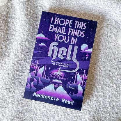 A book titled "I Hope This Email Finds You In Hell" by Mackenzie Reed, featuring a purple and blue sci-fi cover illustration, placed on a white textured blanket.
