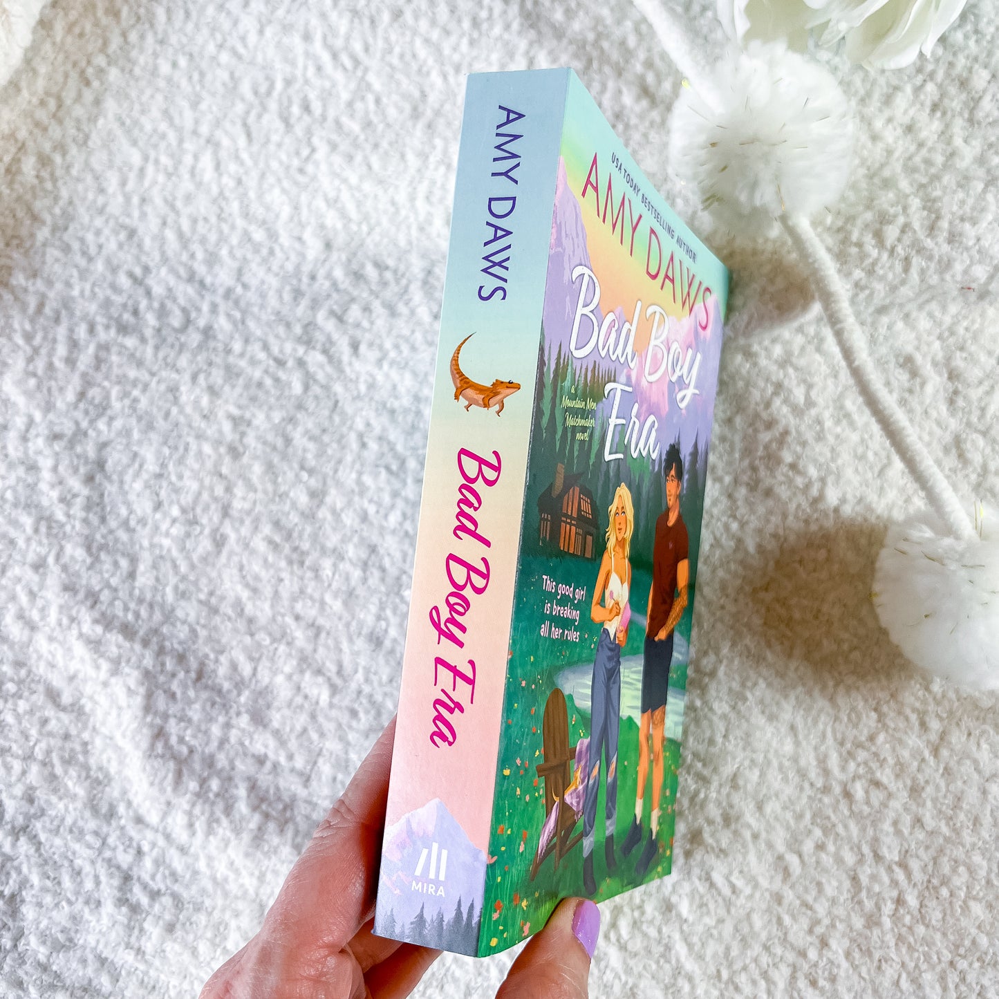 A hand holds a paperback copy of Amy Davis's 'Bad Boy Eve' book, showing the colorful illustrated cover and spine, against a soft white textured blanket background.