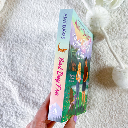 A hand holds a paperback copy of Amy Davis's 'Bad Boy Eve' book, showing the colorful illustrated cover and spine, against a soft white textured blanket background.