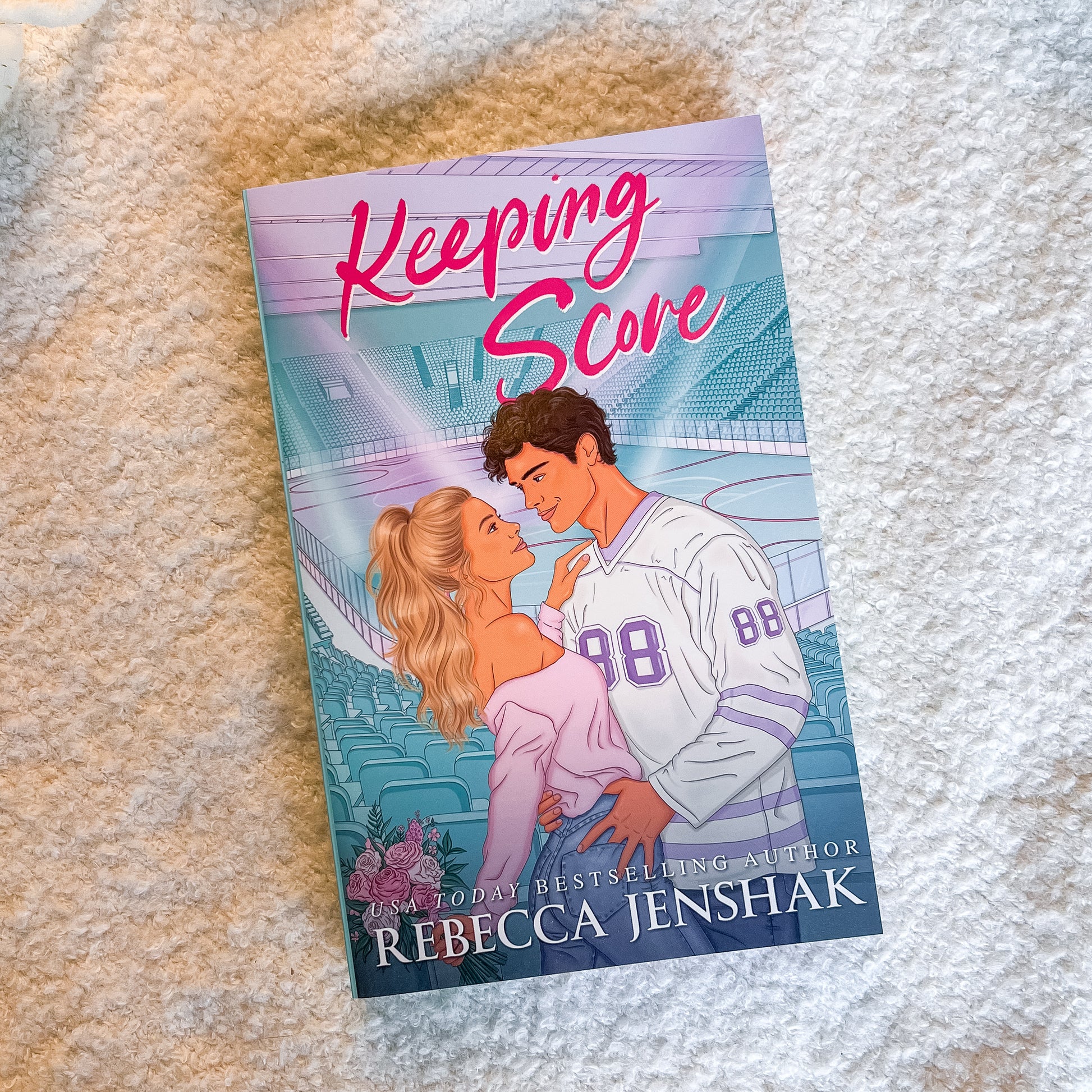 A book titled 'Keeping Score' by Rebecca Jenshak, featuring an illustration of a couple on a sports field, placed on a textured beige fabric background.