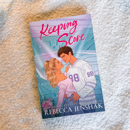 A book titled 'Keeping Score' by Rebecca Jenshak, featuring an illustration of a couple on a sports field, placed on a textured beige fabric background.