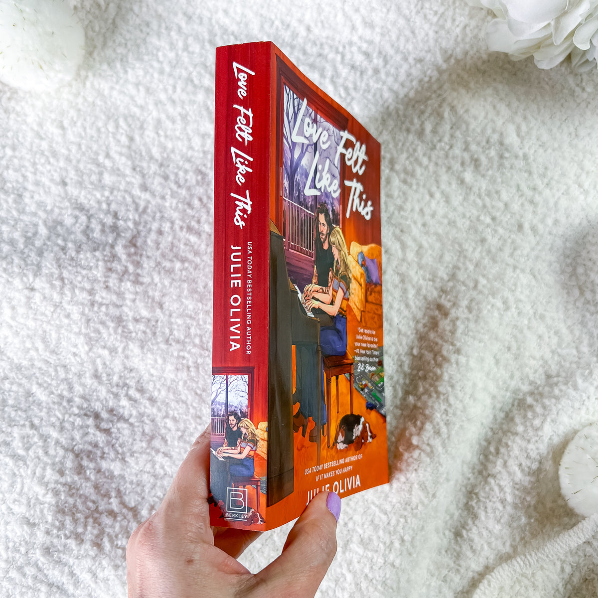 A hand holds a paperback book titled "Love Felt Like This" by Julie Oliva, showing both the red spine and the orange-toned illustrated cover, set against a soft white textured blanket background.