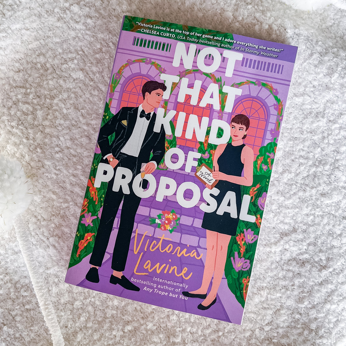 The book cover of 'Not That Kind of Proposal' by Victoria Lavine, featuring an illustration of a man in a black tuxedo and a woman in a black dress against a purple arched doorway background, placed on a textured white fabric surface.