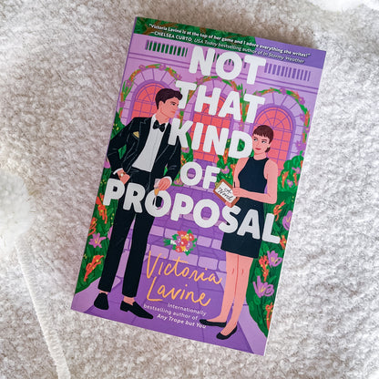 The book cover of 'Not That Kind of Proposal' by Victoria Lavine, featuring an illustration of a man in a black tuxedo and a woman in a black dress against a purple arched doorway background, placed on a textured white fabric surface.