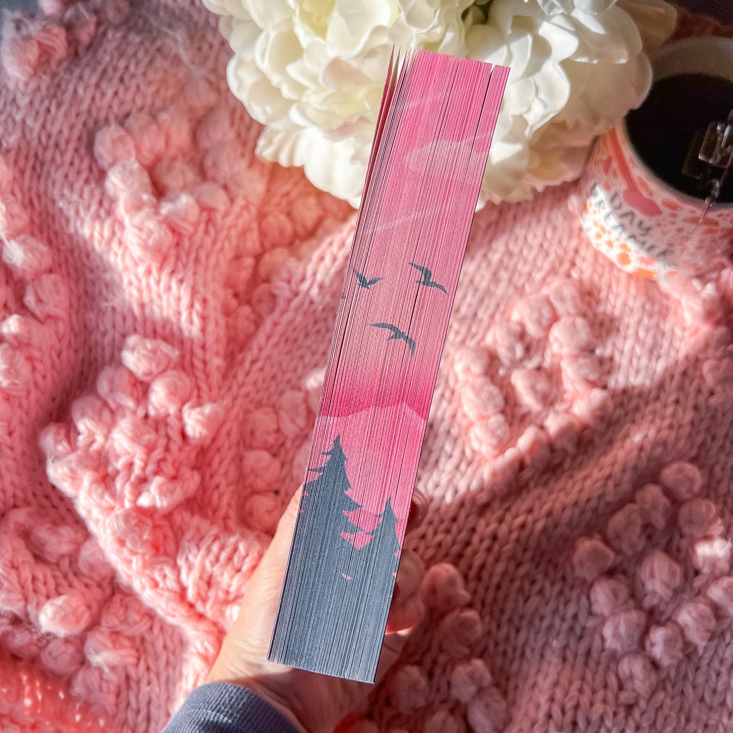 Hand holding a book with floral design page edges on a pink textured surface with flowers and a cup.