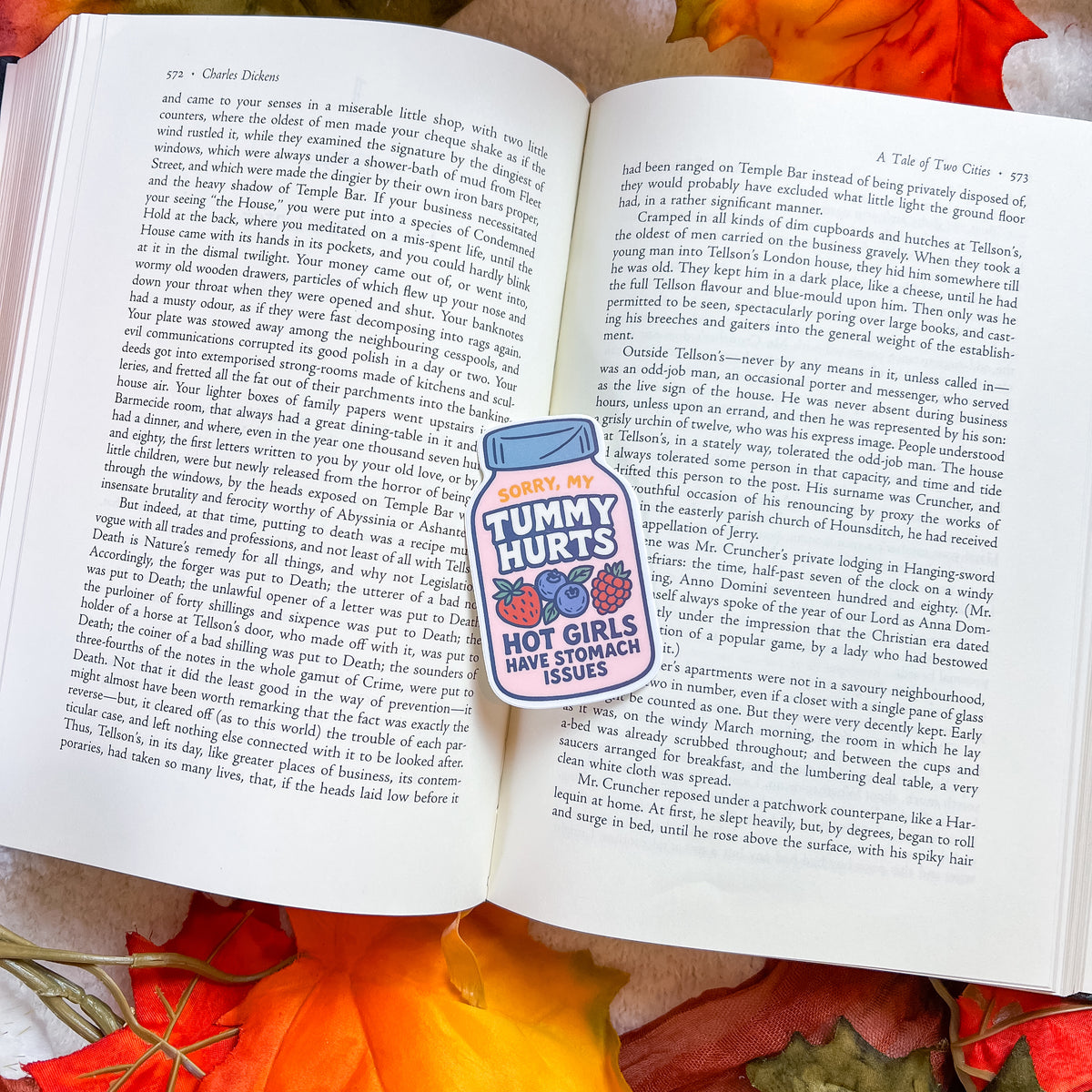 An open book spread with printed text pages, featuring a small sticker on the left page that reads 'TODAY, MY TUMMY HURTS HOT GUY STAMPKICKS ISSUES' with a pink heart and a thumbs-up graphic. The book rests on a red patterned fabric background.