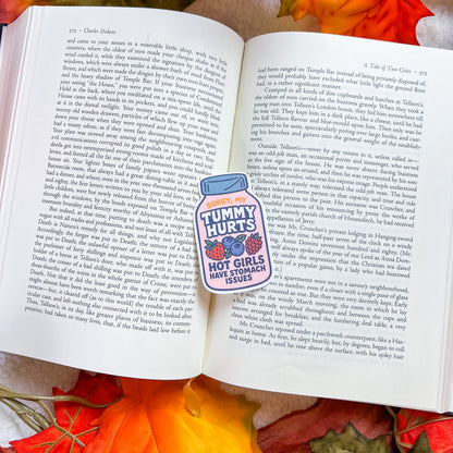 An open book spread with printed text pages, featuring a small sticker on the left page that reads 'TODAY, MY TUMMY HURTS HOT GUY STAMPKICKS ISSUES' with a pink heart and a thumbs-up graphic. The book rests on a red patterned fabric background.