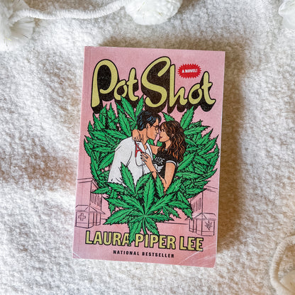 A paperback copy of the novel 'Pot Shot' by Laura Piper Lee lies flat on a white textured blanket. The cover features a pink background with a cartoon illustration of a couple surrounded by green marijuana leaves.