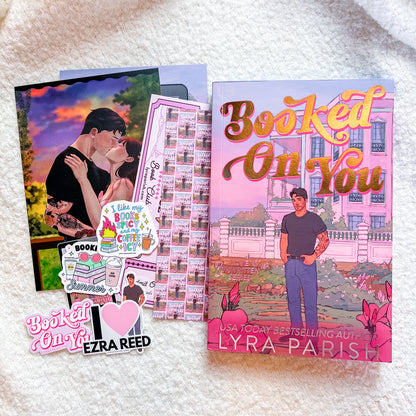 A book-themed flat lay featuring the book cover for 'Booked On You' by Lyra Paris, a matching illustrated card, a sheet of bookmarks, and two stickers on a white textured blanket background.