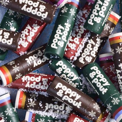 A close-up, top-down view of multiple lip balm tubes arranged on a textured blue surface with white faux snow. The tubes are wrapped in dark brown, dark green, and red paper with white text reading 'poppy & pout' and snowflake graphics.