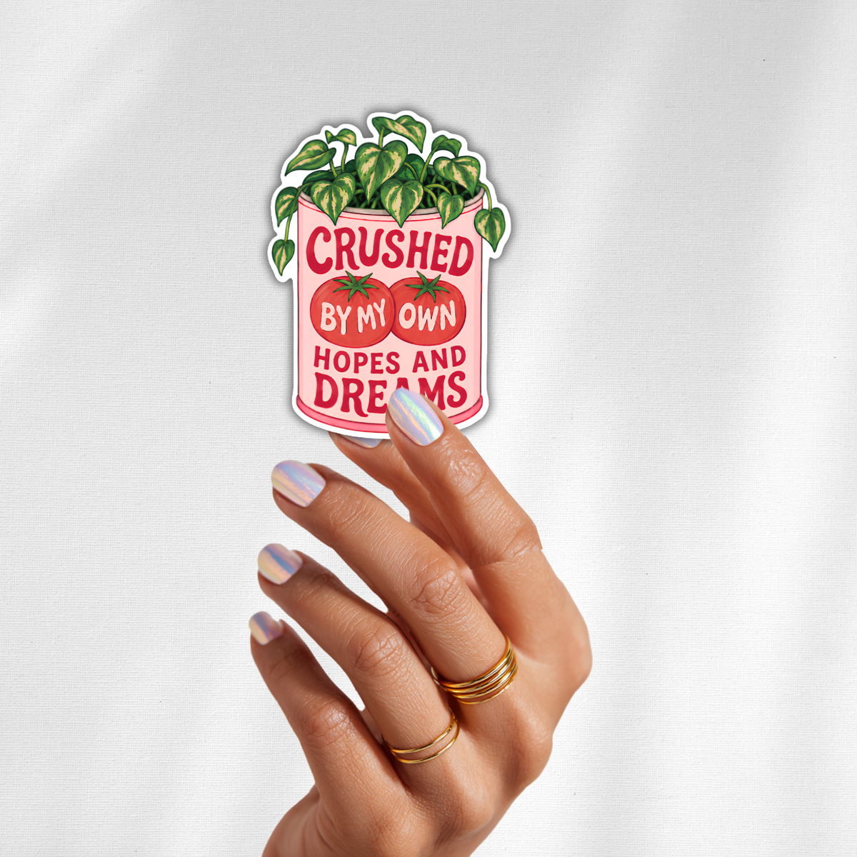 A hand with light purple nail polish and gold rings holds a die-cut sticker featuring a pink tomato can with green leaves on top and the text 'CRUSHED BY MY OWN HOPES AND DREAMS' in red on a white background.