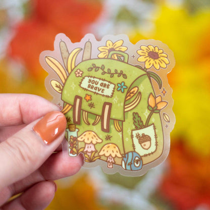 Hand holding a colorful sticker with a green house design against a blurred natural background