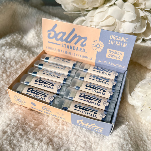An open box of Balm Standard lip balm tubes arranged inside a branded display card, photographed on a soft cream fabric with white flowers in the background. The visible text reads 'balm STANDARD' and 'ORGANIC LIP BALM' with variant notes 'VANILLA BEAN & BLUE CHAMOMILE' and 'WOMAN OWNED.