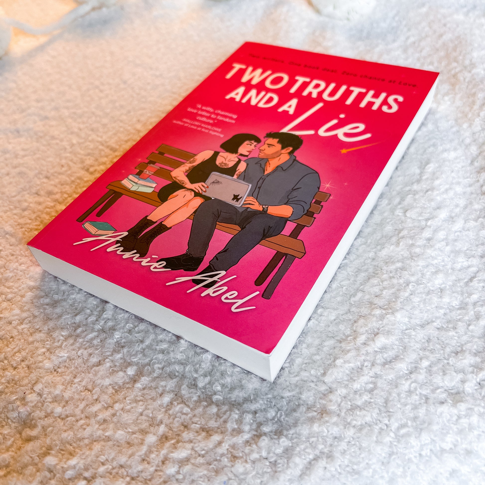 The book 'Two Truths and a Lie' by Amie Abel is shown lying on a textured white blanket. The cover features a bright pink background with an illustration of a couple sitting on a bench using a laptop.