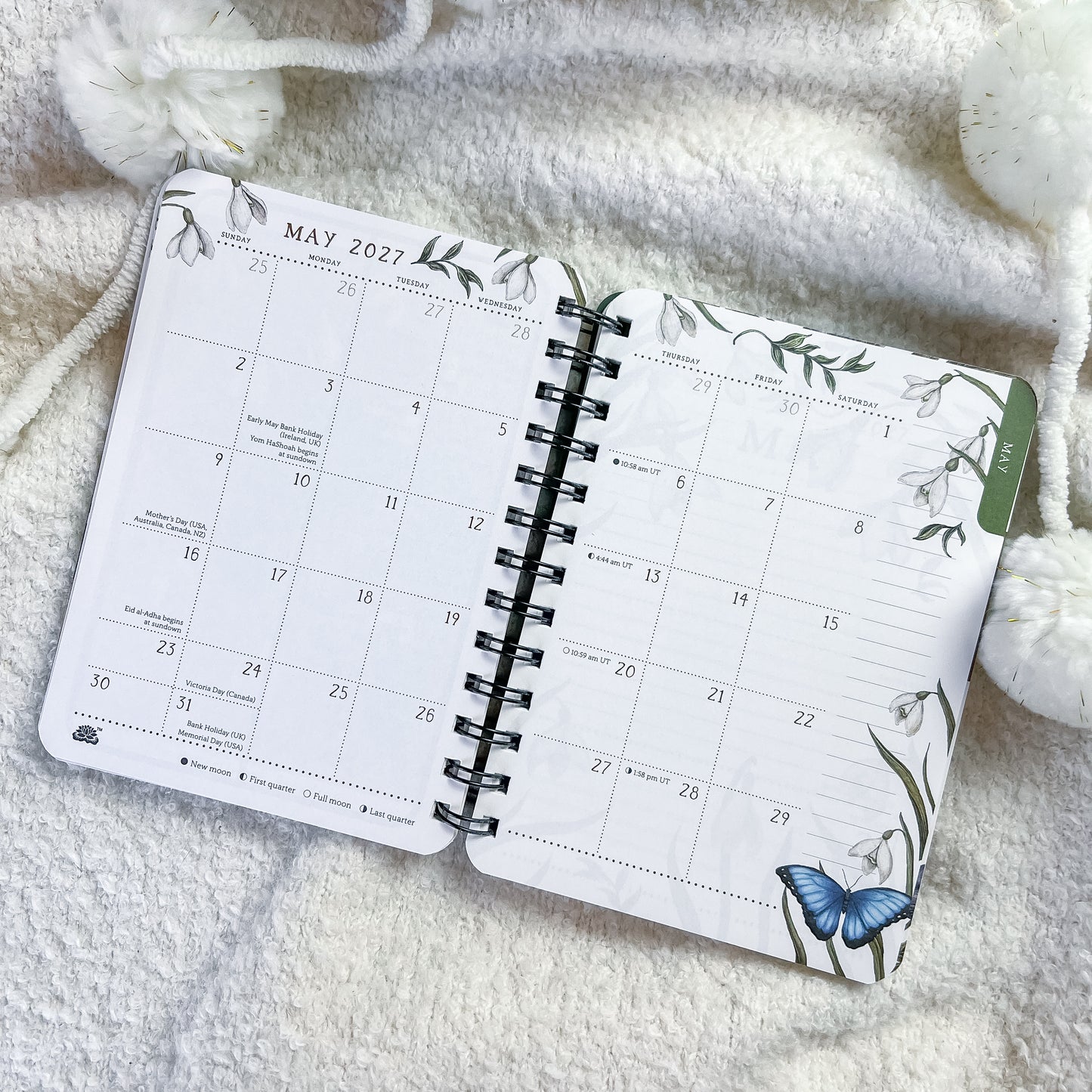 An open spiral-bound planner showing May 2027, styled on a soft white textured blanket with decorative white pom-pom beads.
