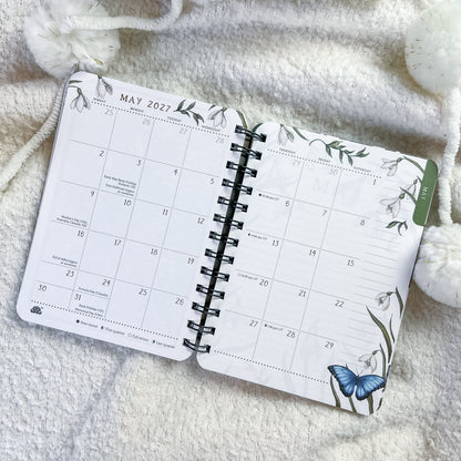 An open spiral-bound planner showing May 2027, styled on a soft white textured blanket with decorative white pom-pom beads.