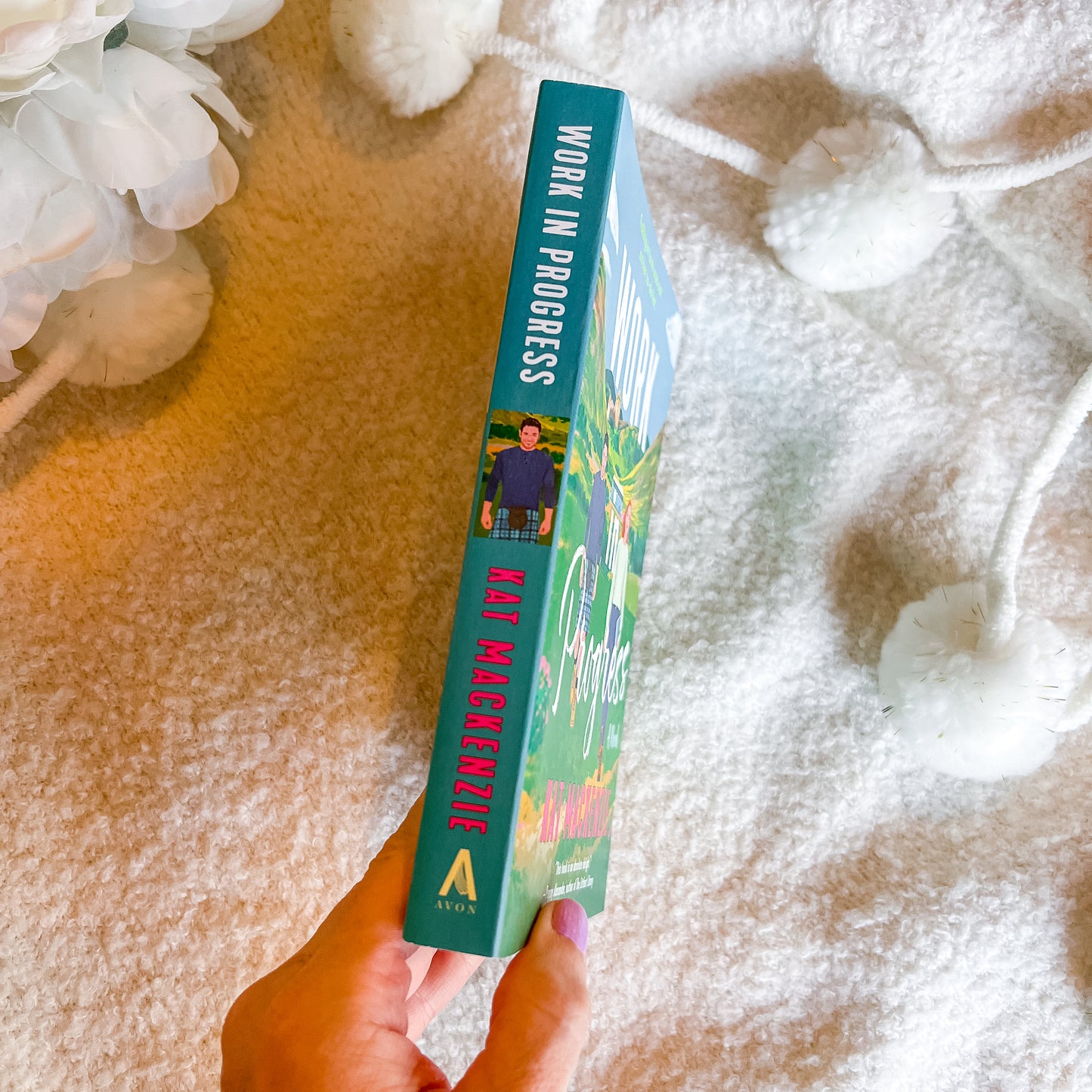 A hand holds a book titled 'Work in Progress', which has a teal spine and colorful cover, set against a cozy white textured blanket with decorative white flowers.