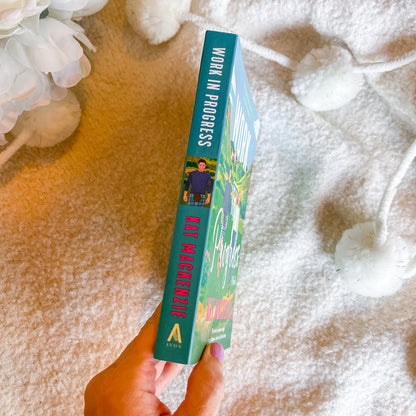 A hand holds a book titled 'Work in Progress', which has a teal spine and colorful cover, set against a cozy white textured blanket with decorative white flowers.