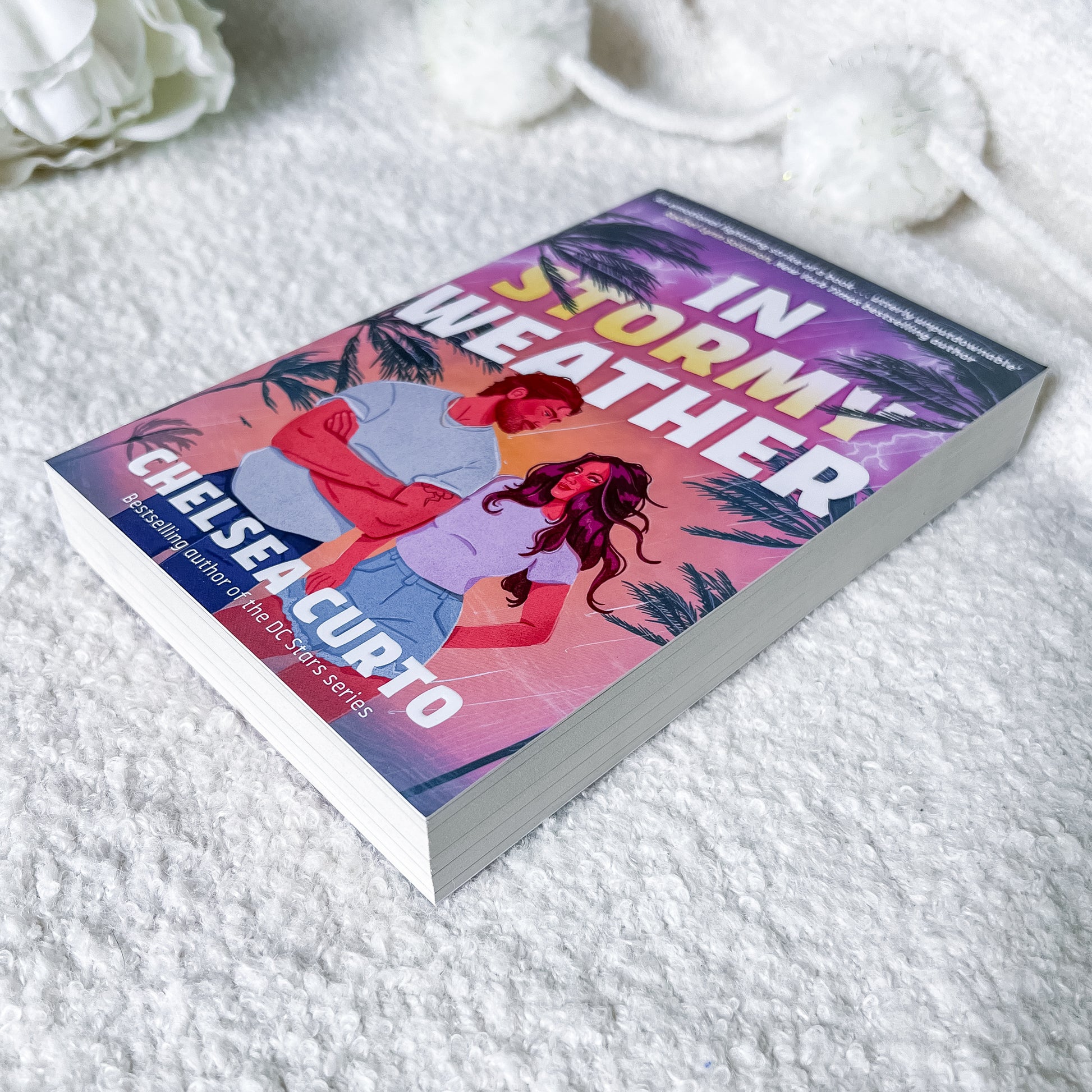 A paperback copy of the book 'In Stormy Weather' by Chelsea Curto lies on a white textured blanket with soft white decorative flowers in the background. The cover features a colorful illustration of a couple under a purple stormy sky with palm trees.