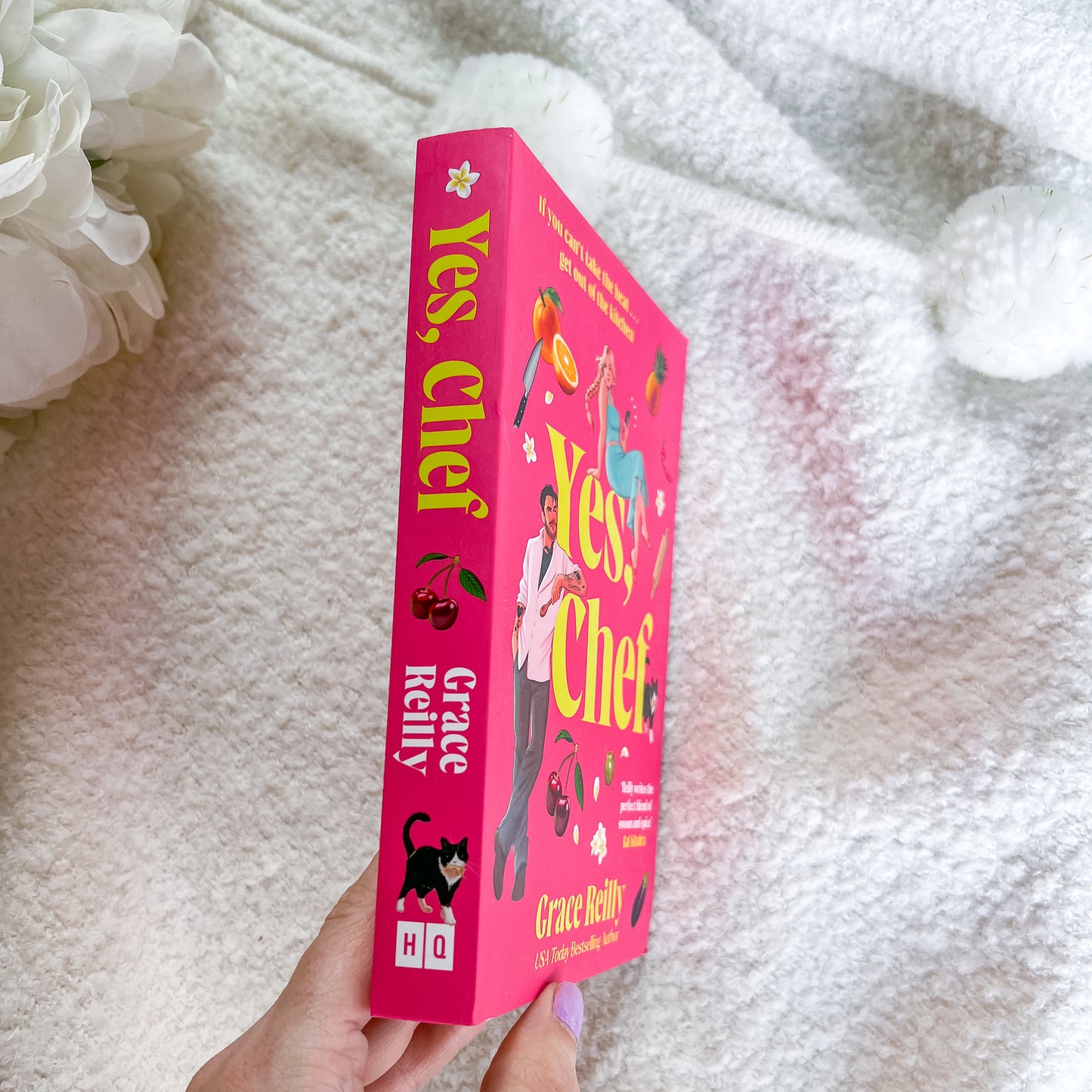 A hand holds a bright pink copy of the book 'Yes, Chef?' by Grace Melny, with white fluffy pom-poms and white flowers in the background.