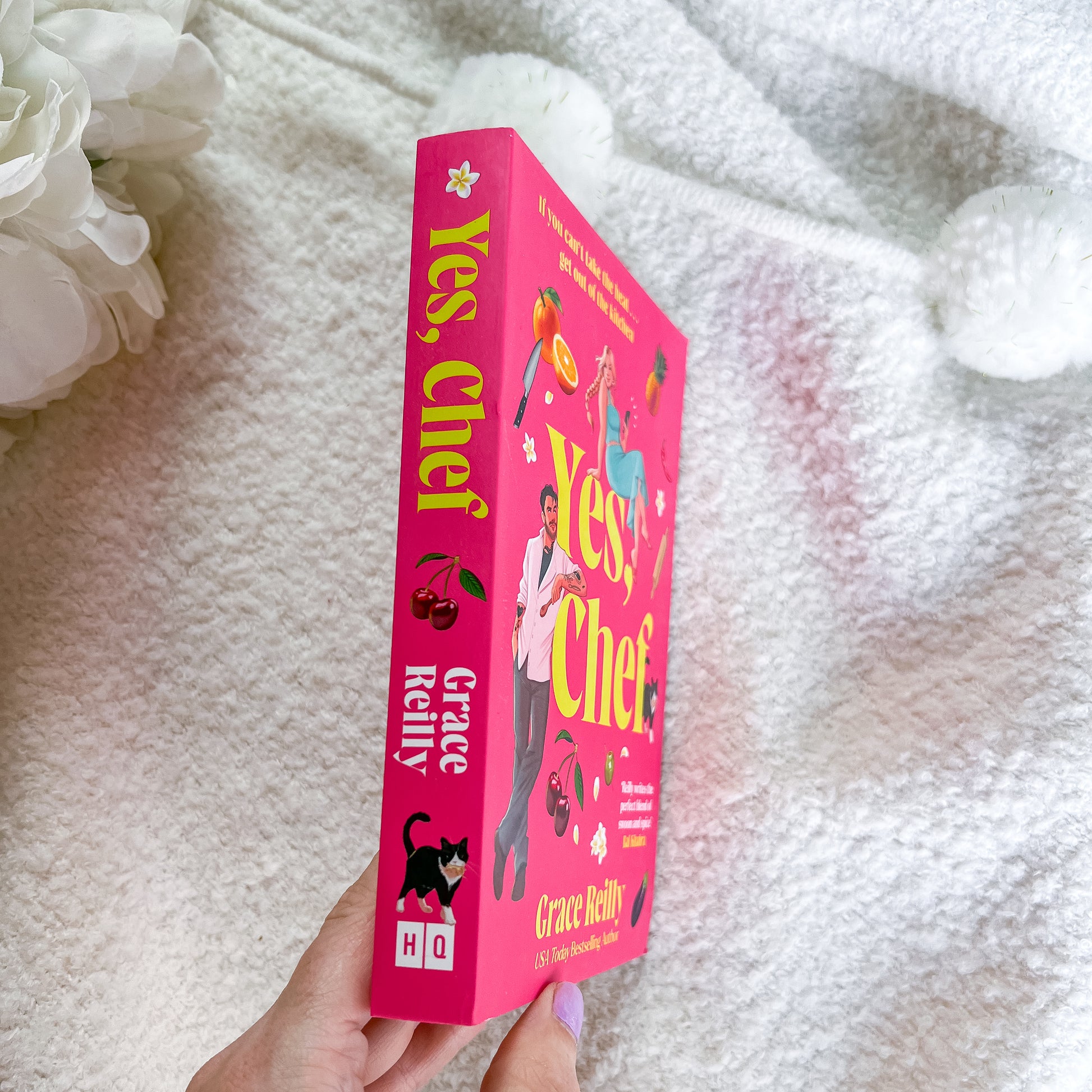 A hand holds a bright pink copy of the book 'Yes, Chef?' by Grace Melny, with white fluffy pom-poms and white flowers in the background.