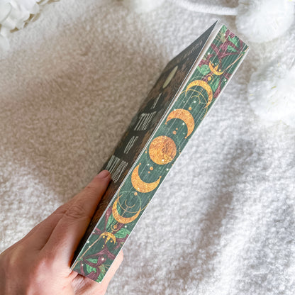 A hand holds a book titled 'The Three-Step Moon' against a soft, textured white blanket background. The book's spine features a detailed design with gold moon phases on a dark background with green and pink botanical patterns.