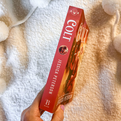 A hand holds a copy of the book by Jessica Peterson, showing the red spine and front cover, set against a soft, textured cream blanket.