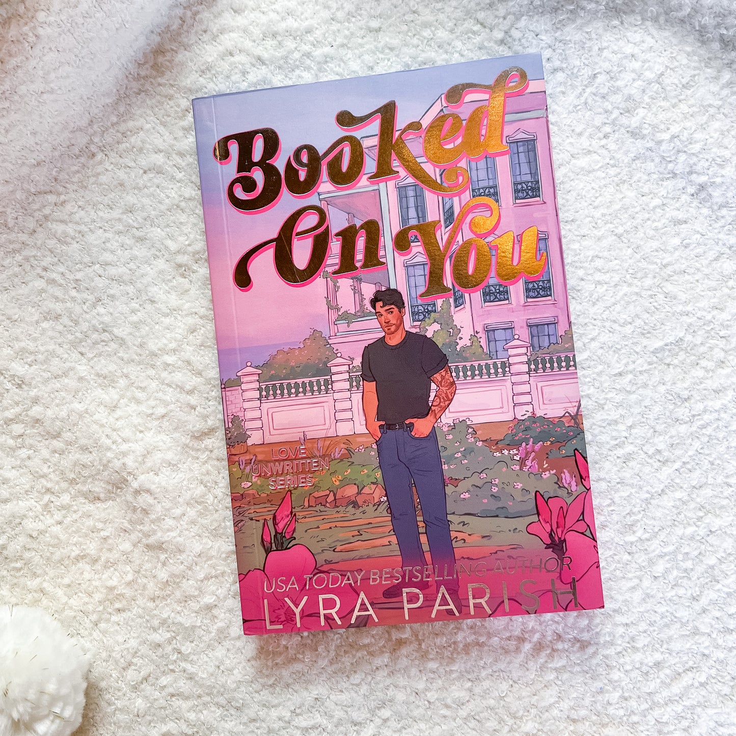 A paperback copy of the book 'Booked On You' by Lyra Parks lies on a soft white textured blanket. The cover features a pink-toned illustration of a woman standing in front of a large pink mansion, with stylized gold and brown lettering for the title.