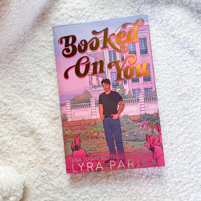 A paperback copy of the book 'Booked On You' by Lyra Parks lies on a soft white textured blanket. The cover features a pink-toned illustration of a woman standing in front of a large pink mansion, with stylized gold and brown lettering for the title.