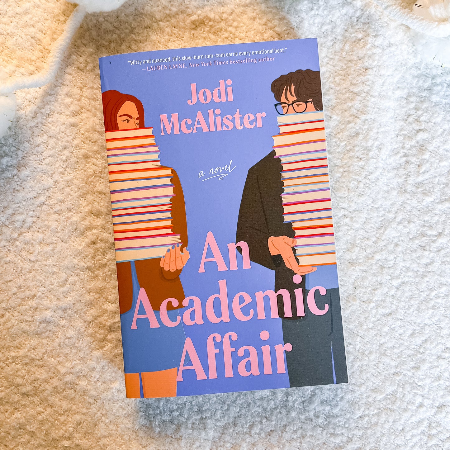 The book cover of 'An Academic Affair' by Jodi McAlister, featuring an illustration of two people covered in books, placed on a textured cream-colored blanket.