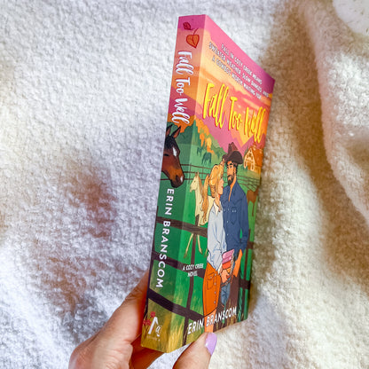 A hand holds a paperback book titled 'Fall Too Tight' by Erin Branson, showing the colorful illustrated cover and spine, set against a soft white textured blanket background.