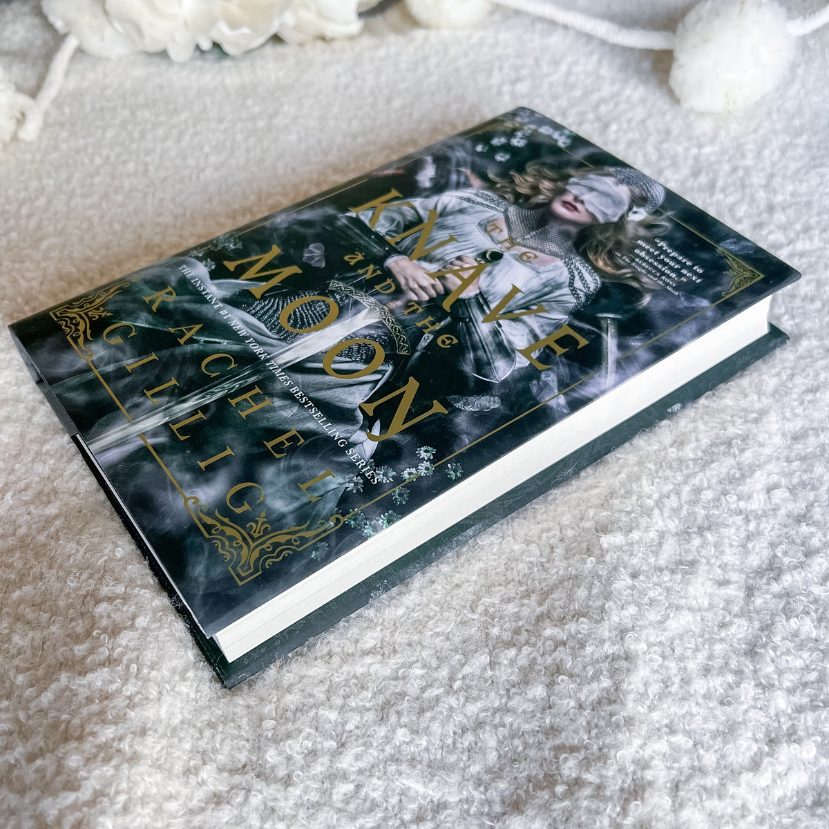 A hardcover book by Rachel Gilligs, featuring a dark gothic cover with gold lettering and an illustration of a blindfolded woman, resting on a textured white carpet.