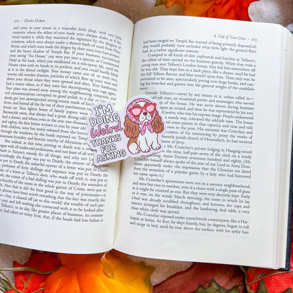 An open book spread showing printed text pages with a colorful cartoon illustration in the center. The illustration shows a girl with long dark hair tied back, wearing a pink and white outfit, with the words 'Doing Weirdo Thanks for Asking' written in a playful font.