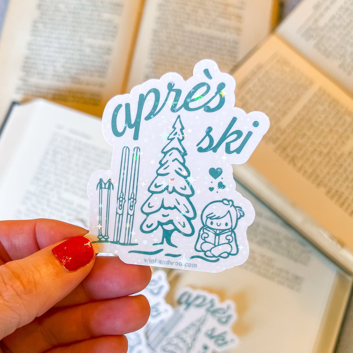 Hand holding a 'apreès ski' sticker with winter-themed illustrations on a blurred background.